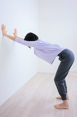 壁に手を付いてお尻を後ろに引き頭からお尻までを伸ばし呼吸する先生